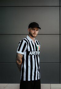 Camisa polo a rayas blancas y negras con cuello, que presenta un logo y la bandera italiana. El modelo también lleva una gorra negra y tiene los brazos tatuados.