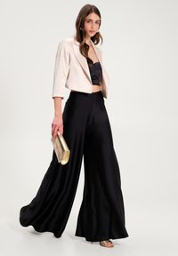 Gekürzte, hellbeige Jacke über einem schwarzen Satin-Top, kombiniert mit weiten schwarzen Hosen. Hält eine goldene Clutch mit subtilen Akzenten.