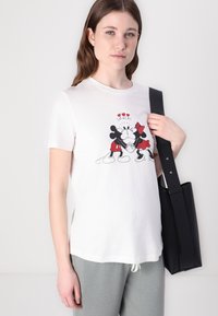 White cotton T-shirt featuring a graphic of Mickey and Minnie Mouse with red accents and "SMACK!" text, paired with gray pants and a black bag.