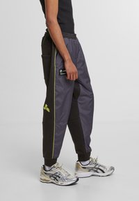 Pilkos ir juodos lengvos sportinės kelnės su geltonais akcentais, elastinga juosmens juosta, šoninės kišenės ir logotipo ženklais prie klubo ir šlaunies.