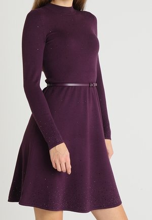 Kobieta ubrana w długą sukienkę z długim rękawem, do kolan, w ciemnym fiolecie, z cienkim paskiem i subtelnymi drobnymi kropkami jako ozdobami.
