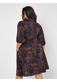 Kleid in mittlerer Länge aus marineblauem Stoff mit einem wirbelnden braunen und orangefarbenen Muster. Hat kurze Ärmel und Seitentaschen. Lässiger Schnitt.