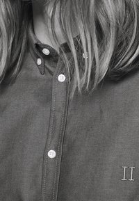 Grijs overhemd met knopen, gemaakt van een textuurstof, witte knopen en een subtiel geborduurd detail met het cijfer "II" rechtsonder.