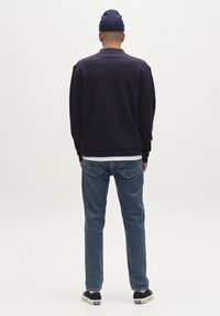 Tema pulover v mornarsko modri barvi z teksturirano površino, nošen čez belo srajco, v kombinaciji s svetlo modrimi kavbojkami in črnimi športnimi čevlji. Na glavi ima volneno kapo.