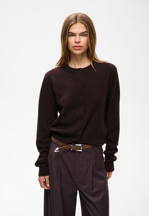 Jeune personne aux cheveux lisses châtain clair porte un cardigan foncé boutonné et un pantalon foncé taille haute avec une ceinture marron.