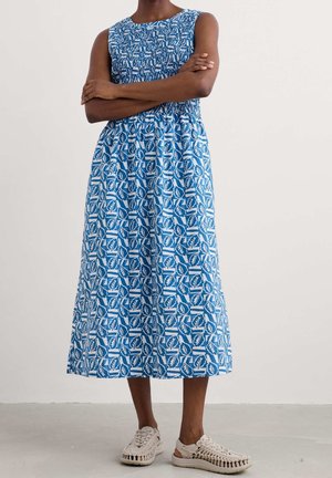Femme les bras croisés portant une robe midi sans manches bleu et blanc à motifs de feuilles et des sandales tissées beiges, debout sur un sol gris.