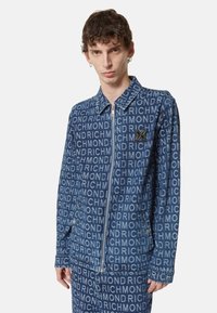 Giubbotto di jeans blu scuro caratterizzato da un motivo con logo "RICHMOND" su tutta la superficie, chiusura frontale con zip, colletto a punta e un accento con marchio.