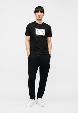 Giovane uomo in piedi con le mani nelle tasche, indossa una t-shirt nera Armani Exchange, pantaloni jogger neri e sneakers bianche su sfondo neutro.