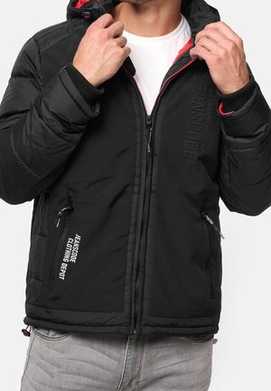 Man wearing black zip-up padded jacket with red lining and "JEANSCODE" branding, adjusting collar, white shirt and grey jeans visible.