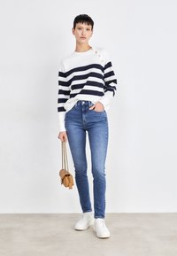 Gestreepte marineblauwe en witte lange mouwtop met schouderknopen, gecombineerd met blauwe skinny jeans. Het model houdt een kleine tan handtas vast en draagt witte sneakers.