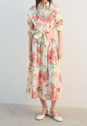 Femme portant une jupe blanche à fleurs et un top à manches longues assorti noué à la taille, associé à des chaussures blanches à talons bas et à bride.