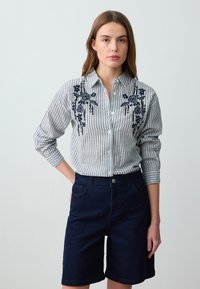 Gestreiftes Hemd in Weiß und Marineblau mit floralem Stickerei auf der Brust, kombiniert mit dunklen Jeansshorts. Knopfverschluss-Design mit umgeschlagenen Ärmeln.