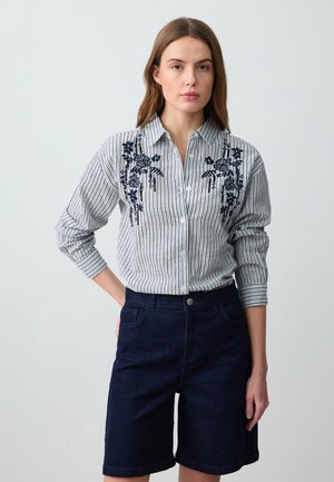 Camicia a righe bianca e blu navy con ricami floreali sul petto, abbinata a pantaloni corti in denim scuro. Design con bottoni e maniche arrotolate.