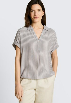 Kobieta ubrana w szarą i białą bluzkę z krótkim rękawem w pionowe paski i beżowe spodnie, stojąca na jednolitym jasnym tle.