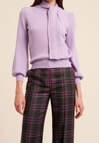 Orchidejový žebrovaný svetr s detailem na zavazování u krku, kombinovaný se šedými károvanými kalhotami s fialovými a modrými linkami, vytvářející texturovaný vrstvený vzhled.