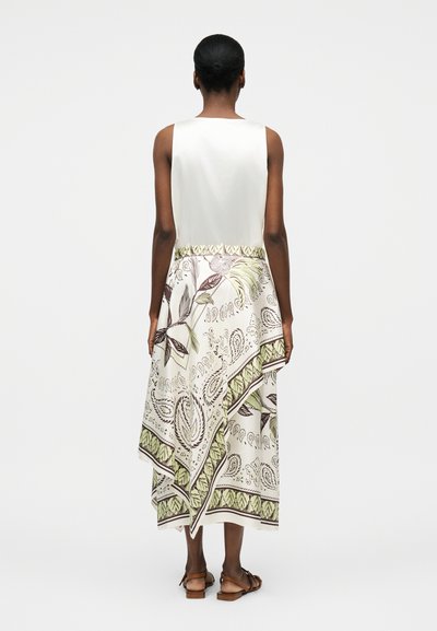 Γυναίκα φοράει λευκό αμάνικο τοπ και ασύμμετρη μίντι φούστα με πράσινο και καφέ φυτικό σχέδιο, εμφανιζόμενη από πίσω, στέκεται σε λευκό φόντο.