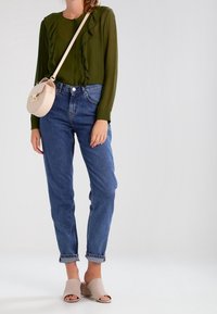 Blusa di chiffon verde scuro con fronte arricciato, jeans azzurri a sigaretta e mule beige; porta una piccola borsa rotonda in rosa chiaro.
