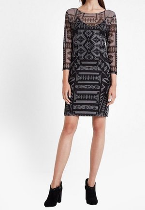 Vestido ajustado en negro y gris con un patrón geométrico, que presenta mangas largas transparentes y un escote redondo, exhibido en una modelo con botas hasta el tobillo.