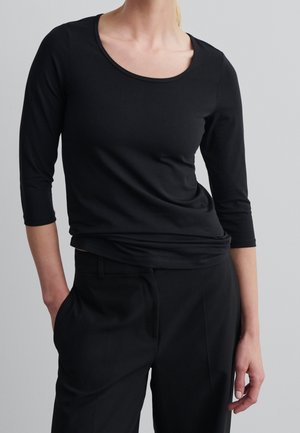 Frau trägt ein figurbetontes, schwarzes Shirt mit U-Ausschnitt und dreiviertel Ärmeln sowie maßgeschneiderte schwarze Hose, steht mit einer Hand in der Hosentasche.