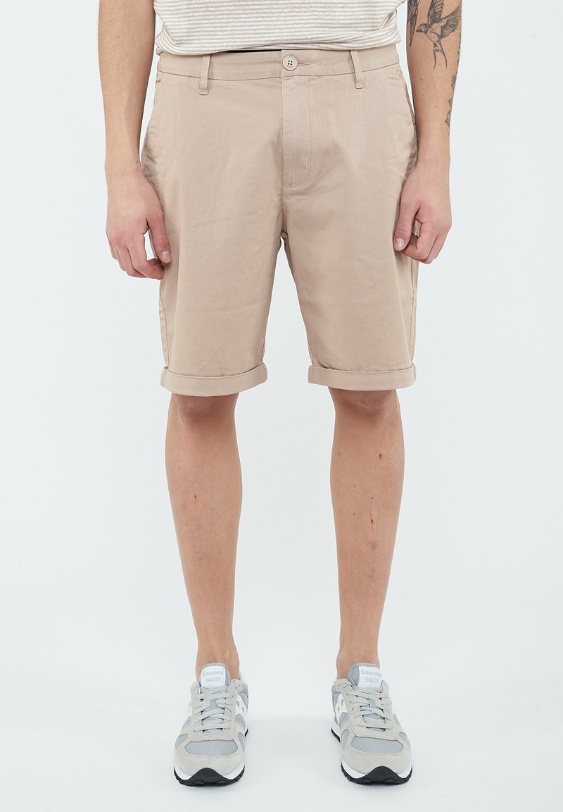 Homme portant un short beige à ourlet retroussé, une chemise blanche à rayures, des baskets grises, avec un tatouage visible sur l'avant-bras gauche.