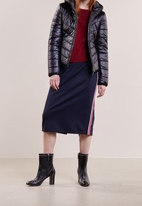 Doudoune noire, pull rouge, jupe midi navy avec des bandes latérales roses, et bottines noires à talons avec des accents cloutés.