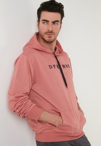 Felpa rosa con tasca frontale, lacci neri e stampa sul tessuto. Realizzata in materiale morbido, presenta polsi ribattuti e una vestibilità casual.