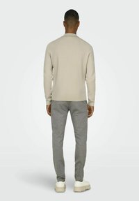 Beige Pullover mit langen Ärmeln, kombiniert mit grauen Jogginghosen. Das Modell trägt weiße Schuhe und wird von hinten vor einem hellen Hintergrund betrachtet.