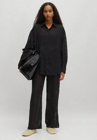 Zwarte doorzichtige blouse met lange mouwen en verticale strepen, gecombineerd met bijpassende gestreepte losse broek. Accessoires omvatten een zwarte handtas.
