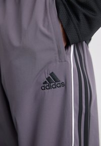 Grå atletiske bukser med sort adidas-logo, sidelommer og sorte striber langs siderne. Fremstillet af glat, letvægtsstof.