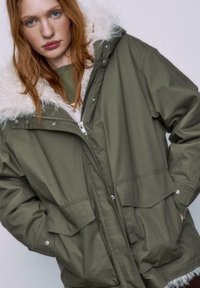 Olivgrüner Parka mit einer Kapuze aus Kunstfell, Reißverschluss vorne und zwei vorderen Taschen. Aus einem glatten, strapazierfähigen Material mit lockerer Passform.