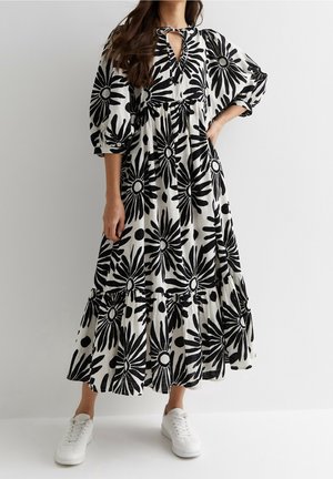 Femme portant une longue robe blanche avec un grand imprimé floral noir, manches bouffantes et baskets blanches, debout avec une main sur la hanche.