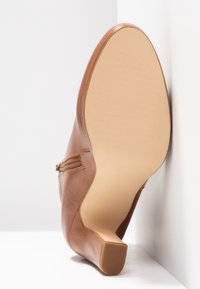Chaussure à talon en cuir marron positionnée sur le côté montrant une semelle beige lisse et un talon carré court sur fond blanc.