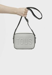 Szara skórzana torba crossbody z wytłoczonym logo "TOUS", prostokątny kształt, gładka tekstura i regulowany czarny pasek.