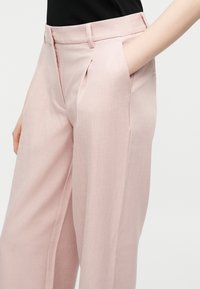 Άτομο που φοράει ροζ παντελόνια ψηλόμεσα με φαρδιά μπατζάκια, πιέτες και πλαϊνές τσέπες, με το χέρι να στηρίζεται μέσα στην δεξιά τσέπη, μαύρο επάνω μέρος εμφανές.