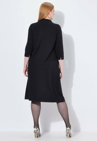 Schwarzes, knielanges Kleid mit Dreiviertelärmeln und seitlichen Schlitzen. Das Modell trägt gemusterte Absatzschuhe und schwarze, transparente Strumpfhosen. Glattes Material.