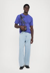 Purple T-shirt with black text, light blue wide-leg jeans, and black shoes. A small black crossbody bag is worn across the body.