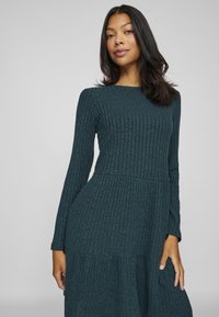 Türkisfarbendes, geripptes Kleid mit langen Ärmeln, rundem Ausschnitt und gestuften Saum. Weicher Stoff mit vertikalen Streifen. Enganliegender Oberteil, der an der Taille ausgestellt ist.