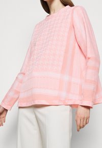 Jasnoróżowa bluzka z długim rękawem o fakturowanym wzorze, z motywem houndstooth i zygzaków, okrągłym dekoltem i luźnym krojem.