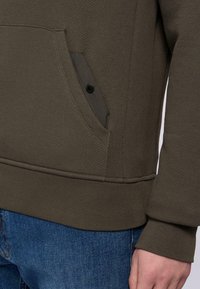 Oliwkowa bluza z kapturem o teksturowanej tkaninie, z boczną kieszenią na napę oraz ribbowymi mankietami. Nosi ją z niebieskimi dżinsami.