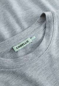 Graues T-Shirt mit rundem Ausschnitt, aus weicher Baumwolle. Mit einem mintgrünen Etikett mit der Aufschrift "CODELLO", das am Kragen genäht ist.