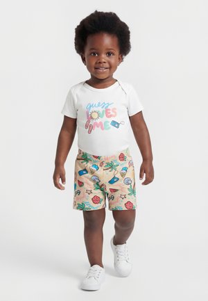 Kleinkind mit lockigem Haar, das ein weißes T-Shirt mit buntem Text und gemusterte Shorts trägt, läuft und lächelt auf einfarbigem Hintergrund.