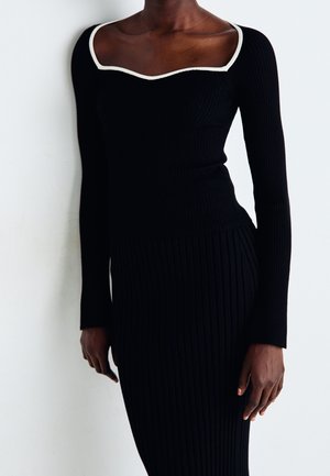 Eine dunkelhäutige Person in einem schwarzen, gerippten langärmeligen Kleid mit quadratischem Ausschnitt und weißem Saum, die vor einem schlichten, hellen Hintergrund steht.