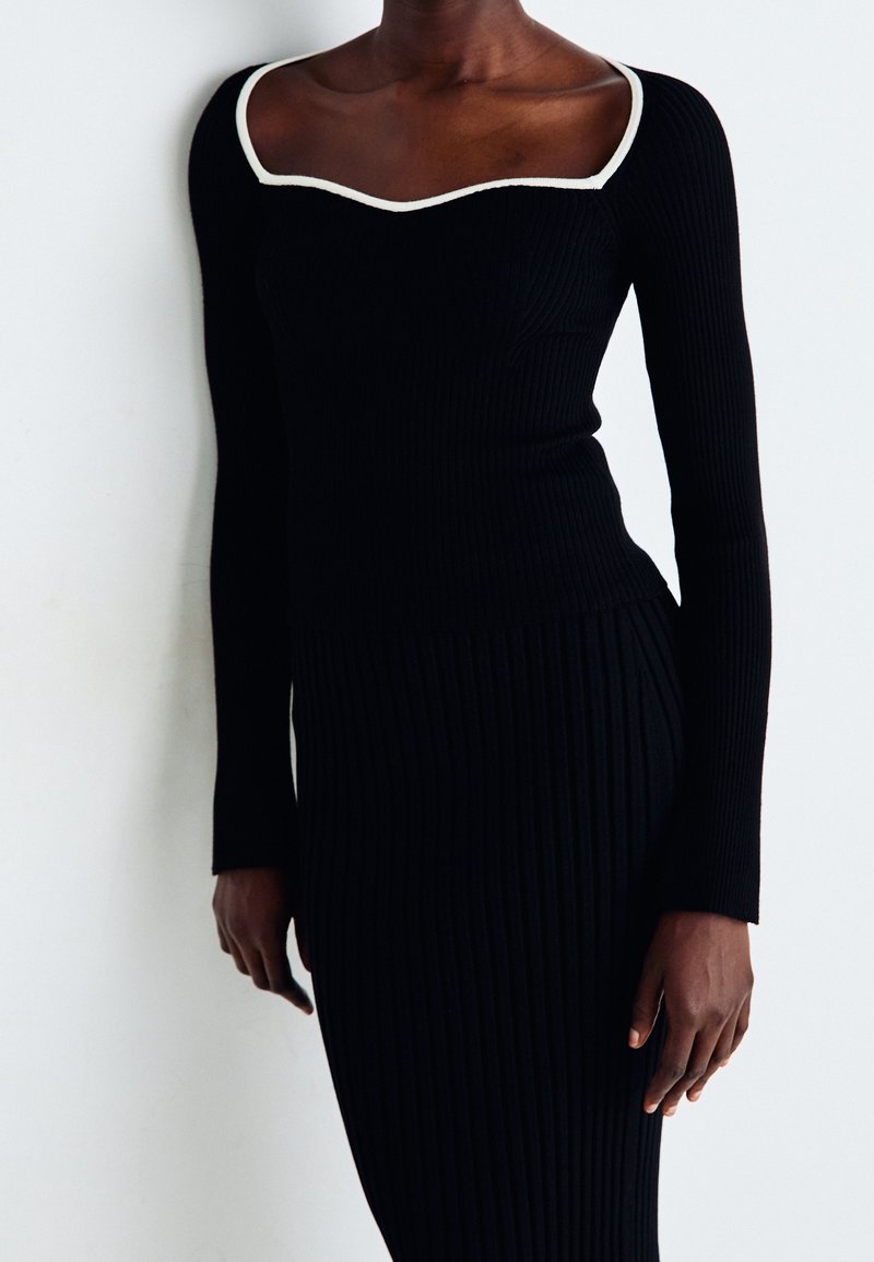 Mörkhyad person i en svart, rätlinjig långärmad klänning med fyrkantig halsringning och vit kant, stående mot en enkel ljus bakgrund.