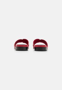 Sandales rouges avec une sangle texturée lisse, arrière ouvert et une semelle plate en caoutchouc noir. Courbure notable dans le design de la sangle au niveau des orteils.