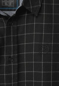 Svart rutigt flanellskjorta med knapp-up design. Har grå rutor, ett upphöjt logobadge och en kontrasterande innervalskrage.