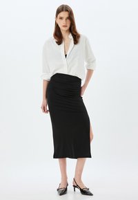Jupe midi noire ajustée avec une fente latérale, associée à une chemise blanche ample à boutons. La tenue présente un design élégant et contemporain.