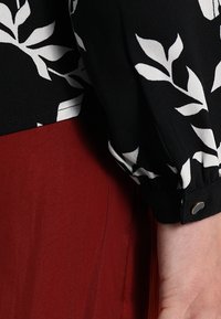 Blusa nera con motivo a foglia bianca, maniche arricciate e dettagli con bottoni. Sotto, è parzialmente visibile un tessuto rosso liscio.