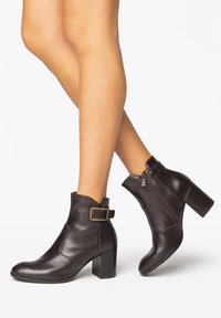 Botas de cuero negras hasta el tobillo con un tacón cuadrado, cremallera lateral y un acento de hebilla en el tobillo. Textura suave y diseño aerodinámico.