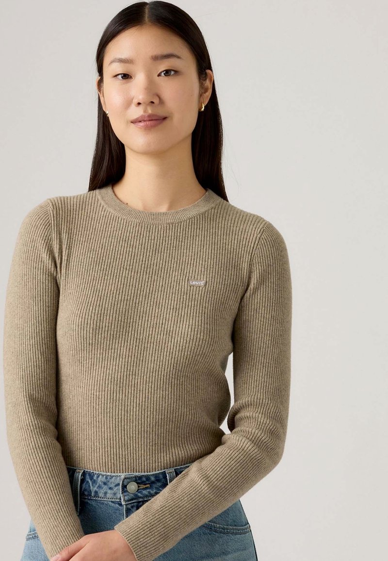 Giovane donna con lunghi capelli scuri che indossa un maglione a coste beige e jeans blu, in piedi contro uno sfondo chiaro e semplice.