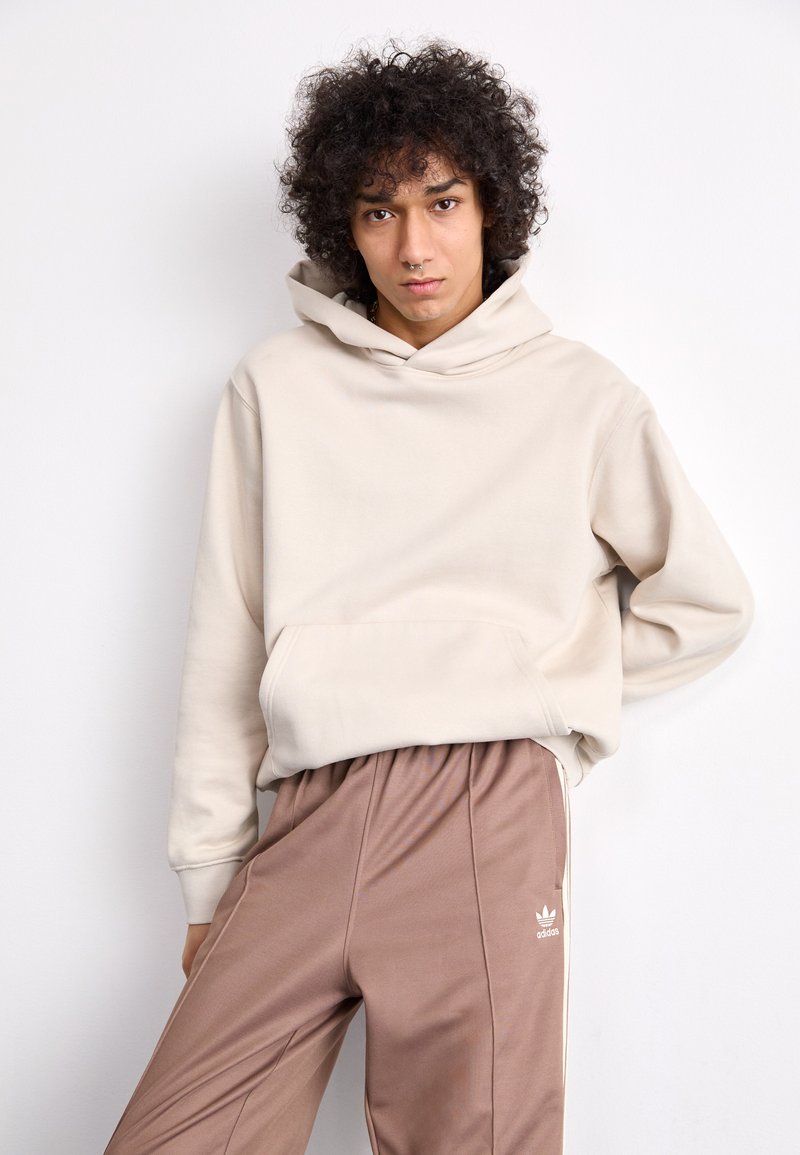 Hoodie beige con tasca frontale e coulisse, abbinato a pantaloni sportivi marroni con strisce bianche ai lati e un piccolo logo.